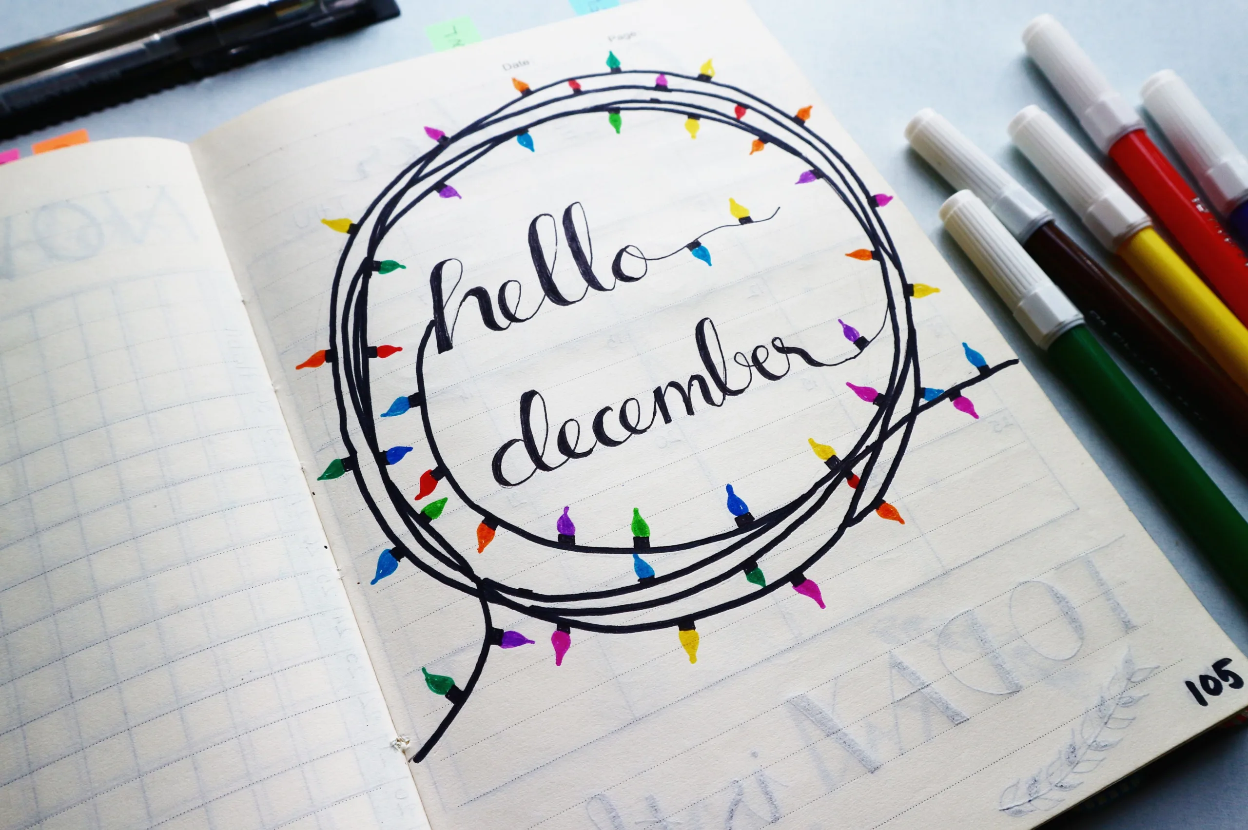 A planner page featuring colourful calligraphy reading 'Hello December' circled by illustration of coloured fairy lights. A collection of coloured pens lay to the side of the page.